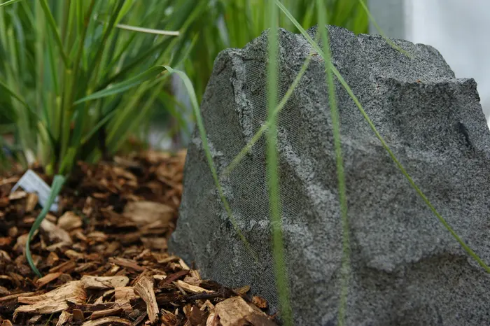 Sonance Rock 63 in Steinform zwischen Gräsern auf Rindenmulch